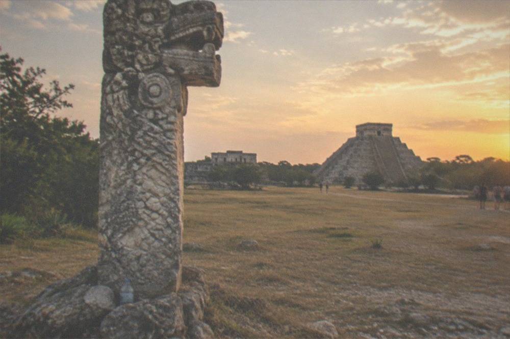 A tall carved stone column standing in an open field with ruins in the distance
