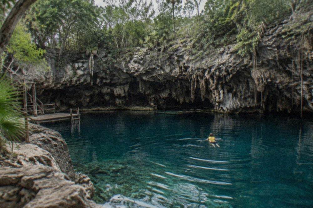 Swimmer floating on still turquoise water in a limestone cenote with rocky walls rising above the surface