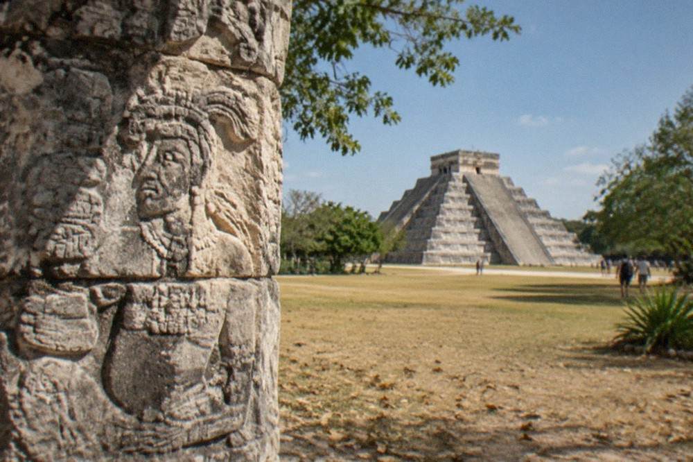 Tall carved stone column of a temple structure at Chichen Itza surrounded by trees and open grass