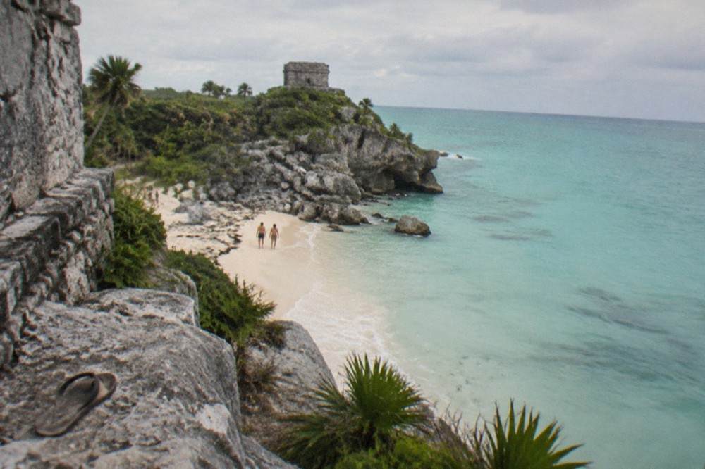 Weathered stone temple walls standing on a cliff edge above a small sandy beach with clear turquoise water