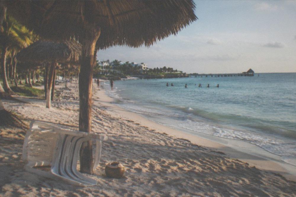 A beach club shoreline with palm trees, thatched umbrellas and calm shallow turquoise water