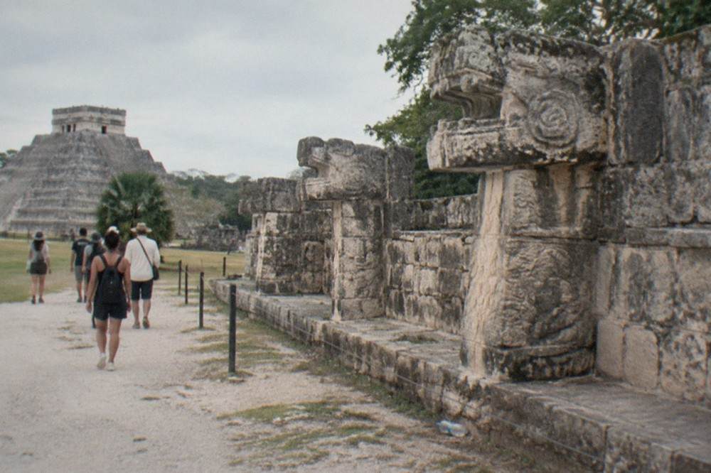 Tour group walking on a stone path beside the carved serpent column ruins at Chichen Itza