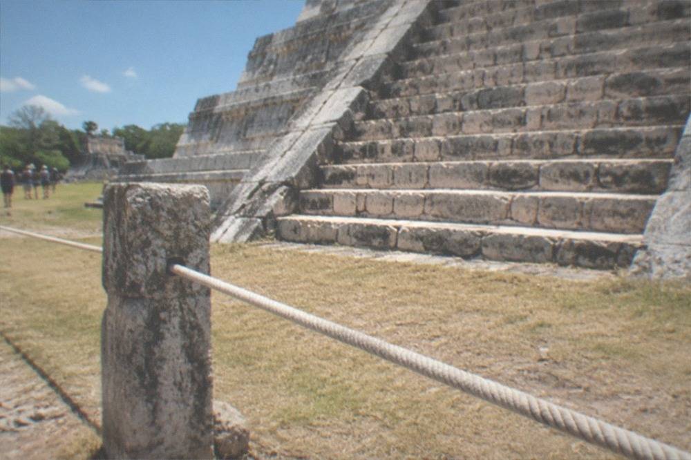 Rope barrier and worn stone steps at the base of a Chichen Itza pyramid, dry grass and clear sky beyond