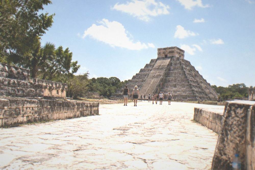 Tourists walking the wide flat stone causeway between ruins at Chichen Itza under midday sun
