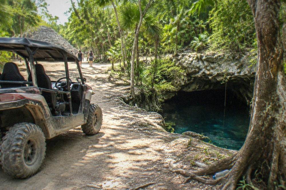 An open buggy parked on a jungle trail beside a rocky cenote entrance