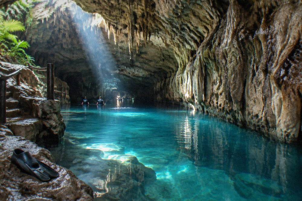 Limestone cave interior with stalactites above still blue water and a shaft of daylight