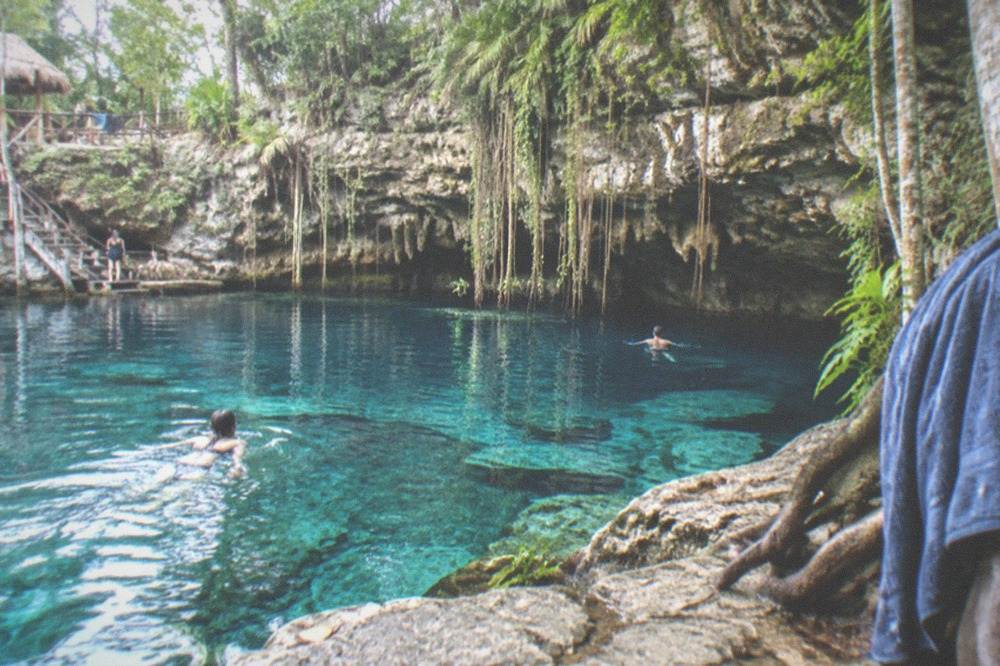 A cenote pool of clear blue water ringed by rock walls and hanging vines in the Riviera Maya jungle